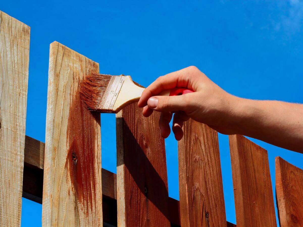A hand stroking a brush of brown paint onto a wooden fence.