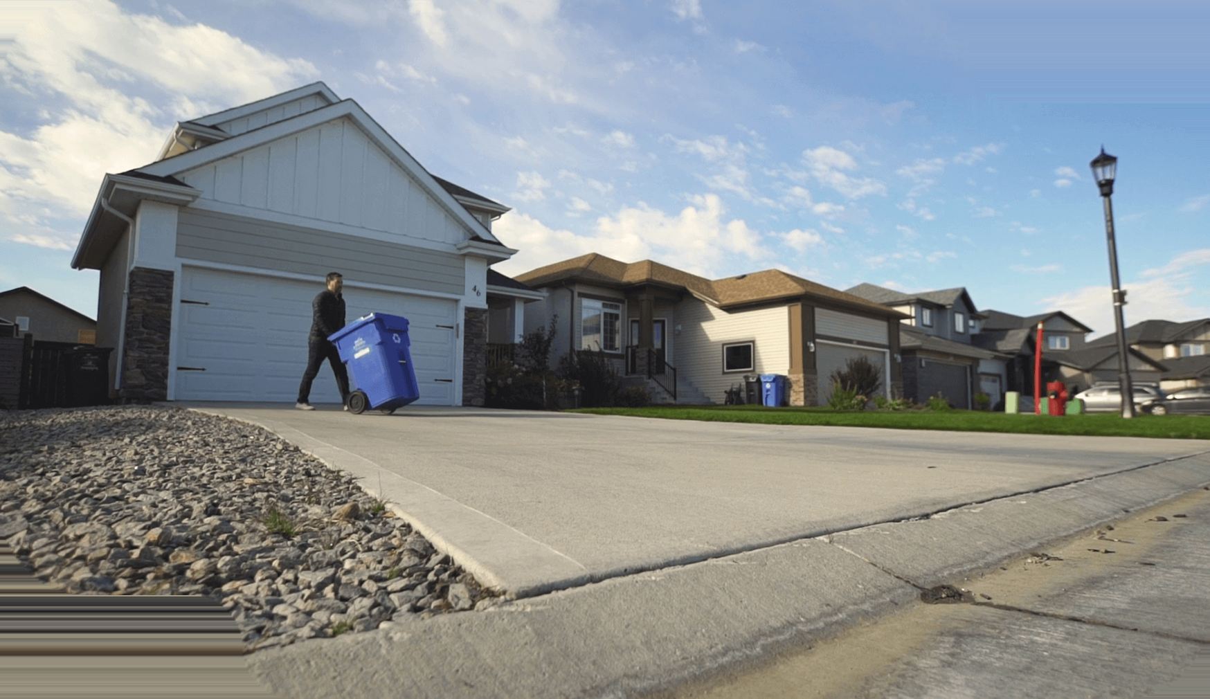 A resident wheeling a blue recycling cart down a suburban driveway